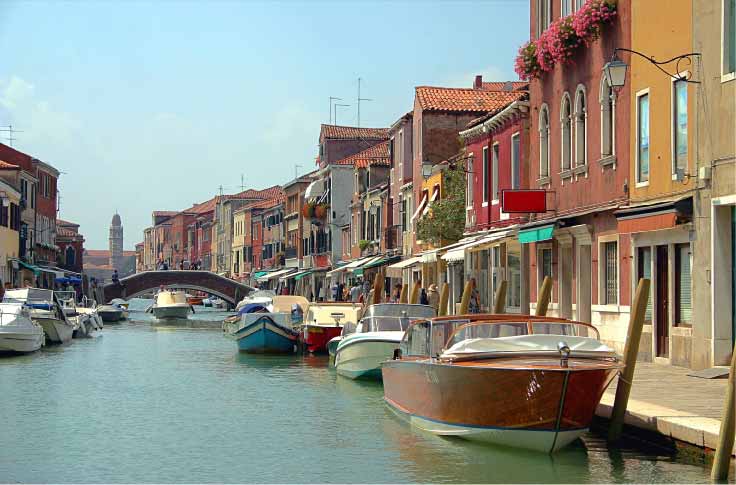 Uma vista panorâmica de um canal em Veneza, Itália, ladeado por edifícios coloridos e barcos atracados. No fundo, uma ponte arqueada atravessa o canal e uma torre de igreja pode ser vista. O céu está azul claro.