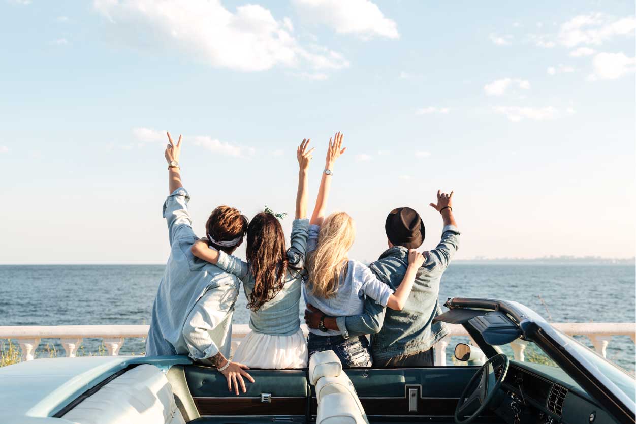 Quatro amigos, de costas, comemorando em um carro conversível próximo ao mar, com os braços levantados.